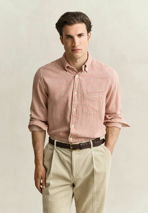 Giovane uomo con capelli castani corti che indossa una camicia rossa a quadri con maniche arrotolate, pantaloni beige in velluto a coste e una cintura intrecciata scura.