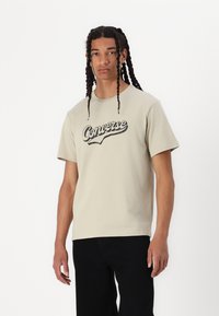 Camiseta de manga corta color beige con un logo en cursiva negro de "Converse" en el pecho, hecha de un suave tejido de algodón. Combinada con pantalones negros.
