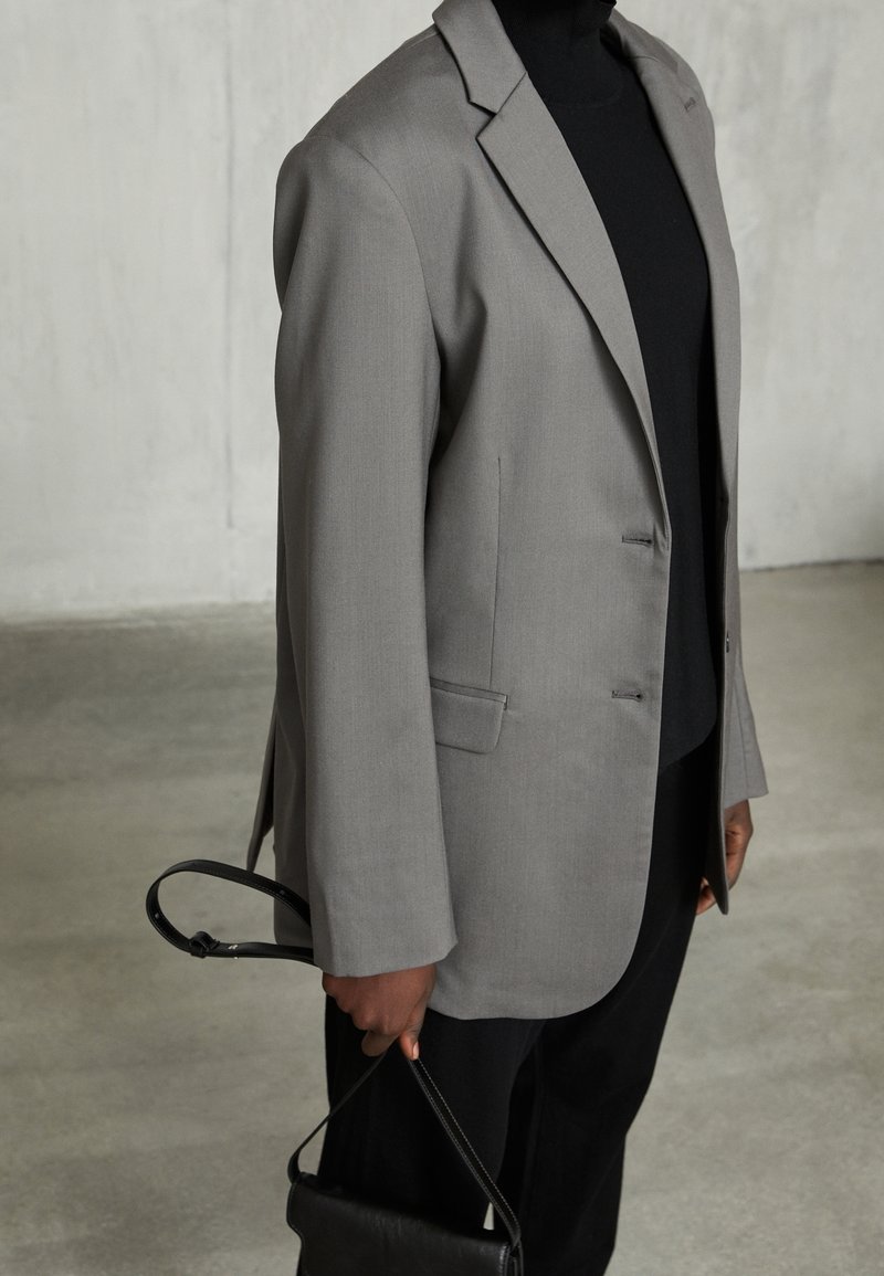 Persoon in een grijze blazer over een zwart top en broek, met een zwarte handtas in de hand aan de strap tegen een vlakke betonnen achtergrond.