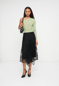 Top de manga longa verde claro com decote assimétrico, combinado com uma saia de renda preta com bainha assimétrica e sapatos de salto alto pretos.