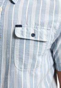 Korte mouwen shirt in lichtblauw met witte verticale strepen. Beschikt over een borstzak met een donkere knop en een klein logolabel op de zak.