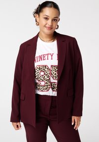 Bordeaux blazer met een enkele knoop, schuine zakken, gelaagd over een wit T-shirt met vetgedrukte rode tekst en een luipaardprint-nummerontwerp.