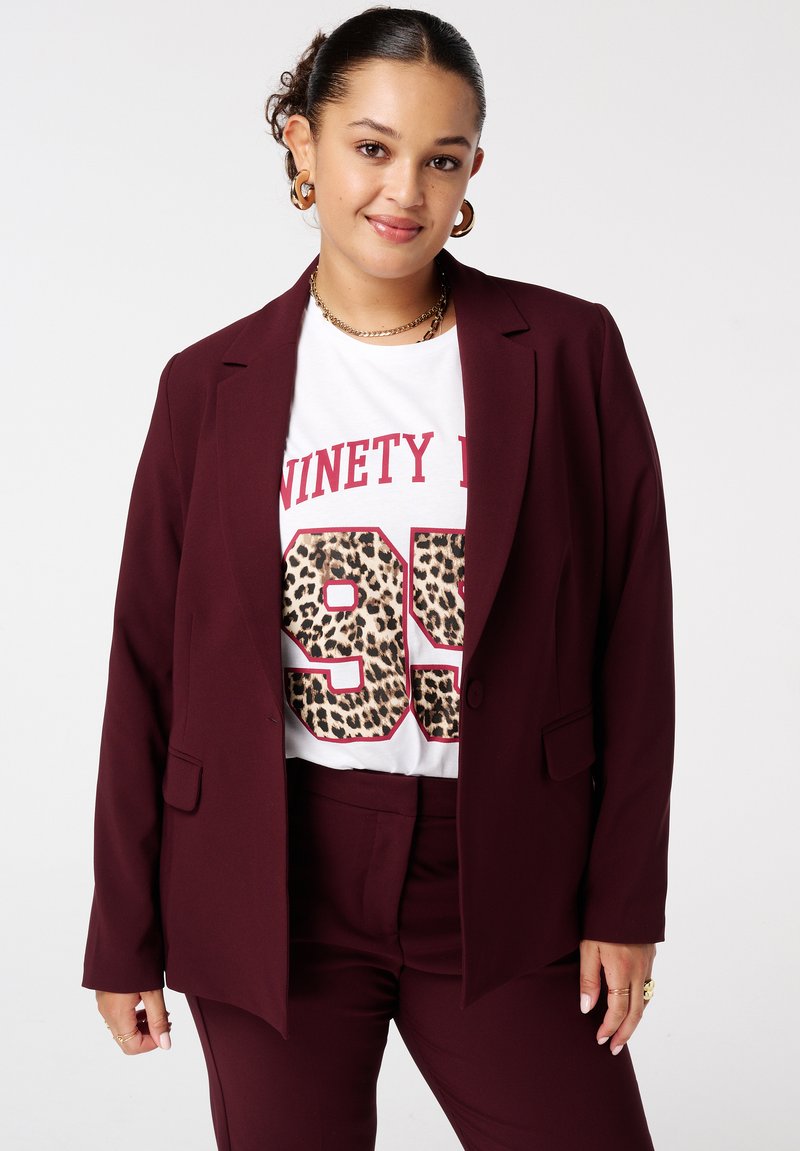 Bordeaux blazer met een enkele knoop, schuine zakken, gelaagd over een wit T-shirt met vetgedrukte rode tekst en een luipaardprint-nummerontwerp.