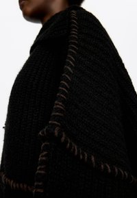 Widok z boku osoby noszącej fakturowany czarny sweter z dużym brązowym pętelkowym szwem wzdłuż szwów na białym tle.