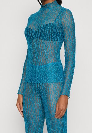 Nainen, joka pukeutunut läpikuultavaan turkoosiin pitsipaitaan ja vastaaviin housuihin, joiden alla näkyy tumma rintaliivit, seisoo yksinkertaisen taustan edessä.