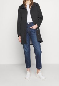 Czarna wodoodporna kurtka z kapturem, zatrzaskami i kieszeniami, noszona na białej koszulce i niebieskich dżinsach, w połączeniu z białymi trampkami.