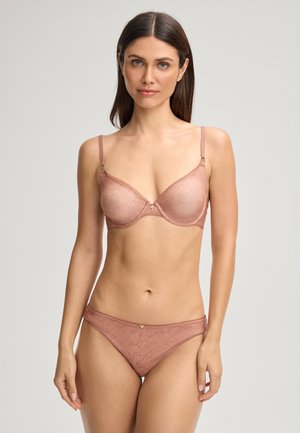 Vrouw met lang donker haar, gekleed in een bijpassende lichtroze beha en slips met bloemenpatroon, staand tegen een eenvoudige neutrale achtergrond.