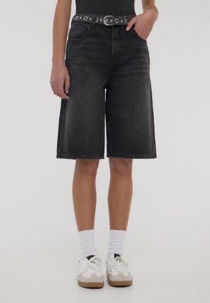 Bermuda shorts in denim nero con gamba ampia. Presentano una cintura con accessori in argento e occhielli. Indossati con calzini bianchi e sneakers chiare.