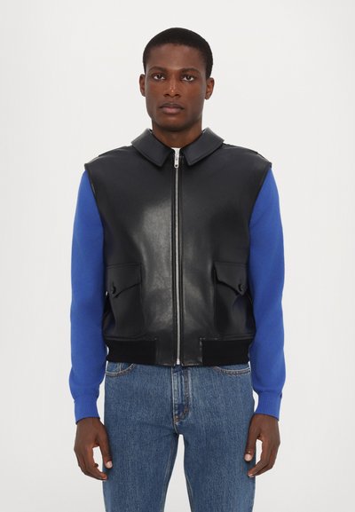 Sort lædervest med høj krave, lynlås foran og to store forlommer, parret med en langærmet blå sweater. Denimjeans fuldender looket.