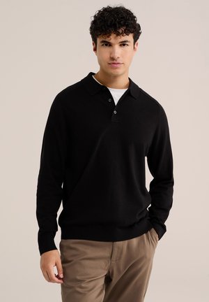Jeune homme aux cheveux bouclés portant un polo noir à manches longues sur un sous-vêtement blanc et un pantalon beige, debout devant un fond uni.