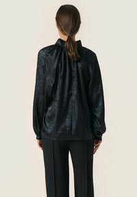 Blusa nera in satin con colletto alto, maniche lunghe e dettagli arricciati sul retro. Texture liscia con un leggero lucido.