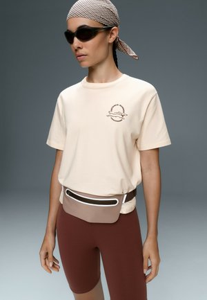 Kvinde iført solbriller, et ternet tørklæde, en beige t-shirt med et løberlogo, brune cykelshorts og en taupe bumbag.