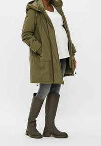 Parka matelassée vert olive avec capuche, poches latérales, associée à des bottes en caoutchouc vert olive montant jusqu'aux genoux. Jean en denim gris en dessous.