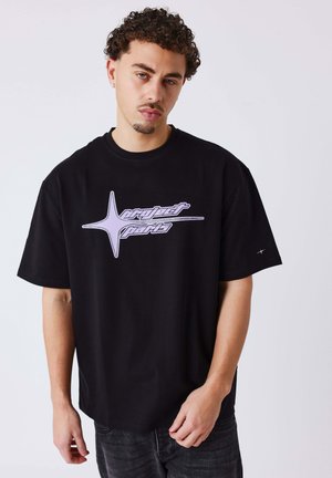Giovane uomo con capelli ricci che indossa una T-shirt nera oversize con una stella viola e la scritta "project parts" sul davanti, in piedi davanti a uno sfondo semplice.