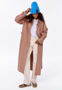 Kobieta poprawiająca niebieską dzianą czapkę, ubrana w długi beżowy płaszcz, biały T-shirt, kremowe spodnie i trampki w panterkę, stojąca na tle białym.