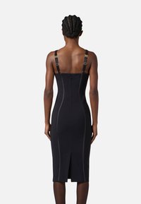 Abito nero aderente con spalline regolabili, dettagli in metallo dorato e zip sul retro. Presenta cuciture a pannello e uno spacco sul retro per facilitare i movimenti.