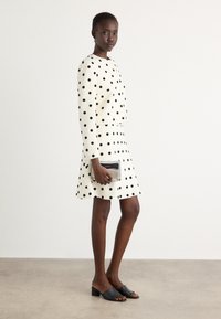 White polka dot dress with long sleeves and flared skirt; paired with a silver clutch and black open-toe mules. Smooth texture, casual design.