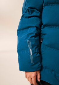 Puffjakke i teal med en struktureret overflade, der har en lynlåslomme og trykte grafikker på ærmet. Nærbillede af armens detalje.