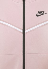 Giacca rosa con zip nera, caratterizzata da strisce diagonali bianche e il logo Nike nero nella parte superiore del petto. Tessuto testurizzato.