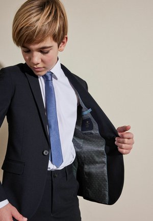 Veste de costume marine avec une texture lisse, un bouton unique et une doublure à pois bleu clair. Portée sur une chemise blanche et une cravate bleue.