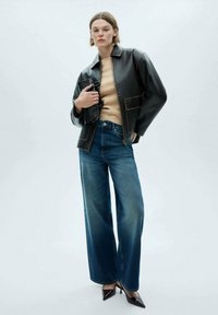 Black leather jacket with a zip, beige sweater, wide-leg blue jeans, and pointed-toe heels, standing against a light gray background.