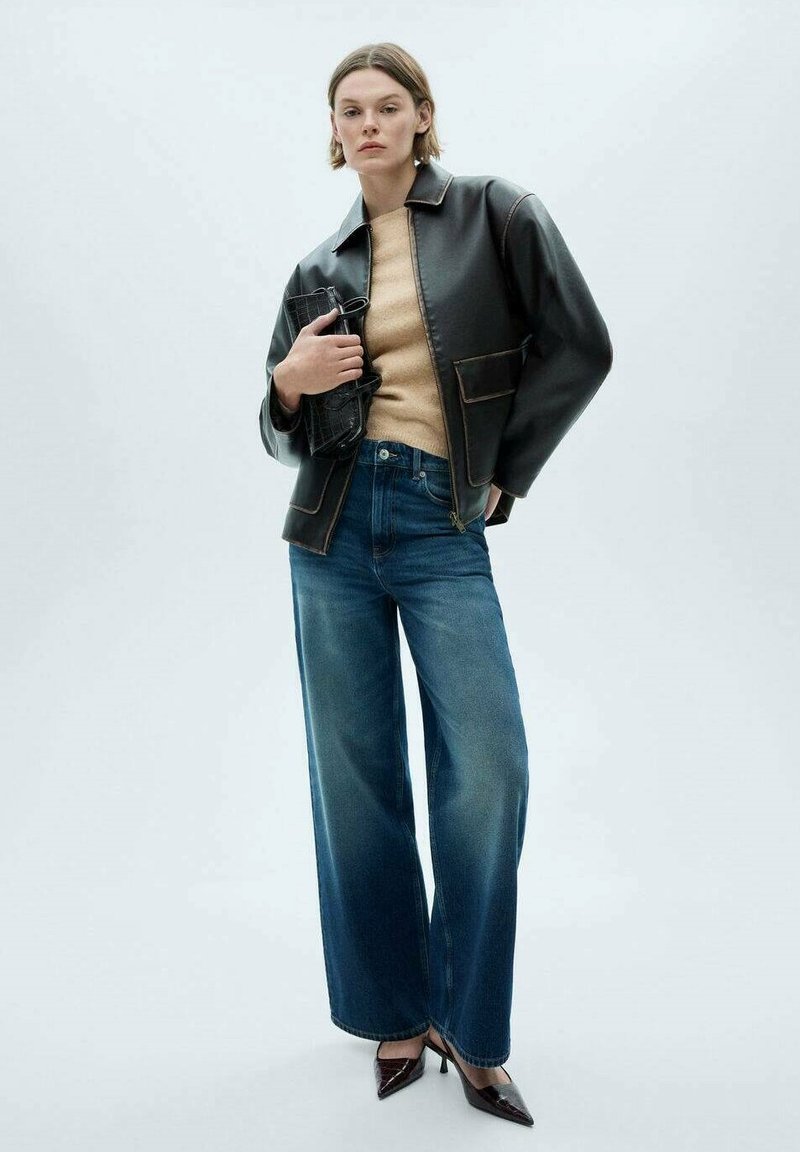 Black leather jacket with a zip, beige sweater, wide-leg blue jeans, and pointed-toe heels, standing against a light gray background.