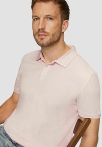 Mann mit kurzem braunem Haar und Bart, der ein hellpinkes Poloshirt trägt und seitlich auf einem Holzstuhl vor einem grauen Hintergrund sitzt.