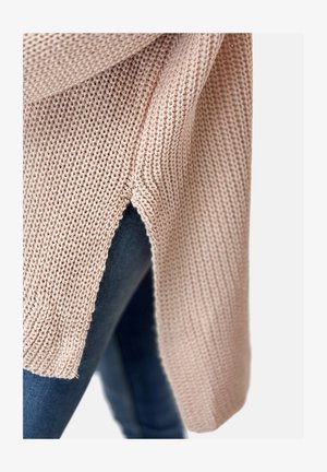 Suéter de punto en suave color rosa con un corte holgado, que presenta una abertura lateral y un tejido texturizado, combinado con denim oscuro.