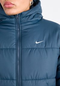 Ναυτικό μπλε μπουφάν με πούπουλα με ψηλό λαιμό, φερμουάρ και μικρό λευκό λογότυπο. Διαθέτει ραμμένες τμήματα και λεία, γυαλιστερή υφή.