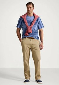 Mężczyzna w niebieskiej, pasek t-shirt, beżowych spodniach, granatowych sneakersach i różowym swetrze zarzuconym na ramionach, stoi z rękami w kieszeniach.