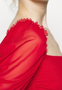 Rotes Kleid mit durchsichtigen, gepufften Ärmeln, die einen Rüschensaum aufweisen. Glatte Textur, taillierte Corsage und gerafftes Detail an der Taille.