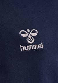 Tessuto blu navy con logo ricamato bianco che presenta un design di ape sopra il testo "hummel". Il materiale ha un aspetto liscio e strutturato.