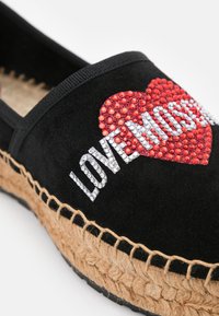 Svart mocka espadrillo med en texturerad jutesula, prydd med röda och silverfärgade rhinestones som formar ett hjärta och texten "LOVE MOST."