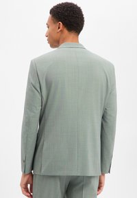 Uomo con indosso un abito grigio chiaro visto di spalle su uno sfondo bianco uniforme.