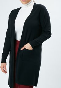 Zwarte cardigan met een open voorkant, geribbelde manchetten en zakken. Zachte stof, lange mouwen en een knie-lange pasvorm over een witte top.