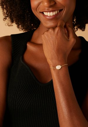 Gouden armband met een gladde, ovale, witte steen in het midden; gedragen om een gebruinde arm tegen een gestructureerd, zwart gebreid topje.