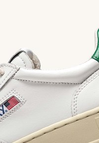 Sneaker bianco in pelle con accentuazione verde sul tallone, suola beige a contrasto e cuciture visibili. Presenta il logo della bandiera americana sul lato.