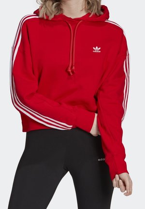 Osoba ubrana w czerwoną bluzę z kapturem Adidas z białymi paskami na rękawach, zestawioną z czarnymi legginsami Adidas.