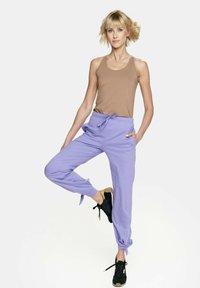 Fioletowe, lekkie spodnie dresowe z elastycznym paskiem w talii, bocznymi kieszeniami i troczkami przy kostkach, stylizowane z beżowym topem na ramiączkach i czarnymi sneakerami.