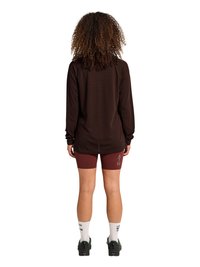Person mit lockigem Haar steht mit dem Rücken zur Kamera, trägt ein schwarzes Langarmoberteil, bordeauxrote Shorts mit dem Schriftzug "ATW", weiße Socken und schwarze Schuhe.