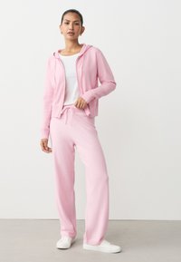 Jasnoróżowa bluza z kapturem na suwak z przodu i ściągaczami przy mankietach, zestawiona z dopasowanymi różowymi spodniami dresowymi z elastycznym pasem. Białe sneakersy.