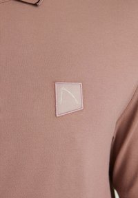 Svetloružová polo košeľa vyrobená z textúrovanej látky, s malým vyšívaným trojuholníkovým logom na nášivke. Krátky golier, ležérny dizajn.