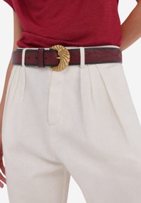 Bruine leren riem met een gestructureerde goudkleurige gesp; gedragen over witte geplooide broeken. Gedetailleerde stiksels op de riem zorgen voor visuele interesse.
