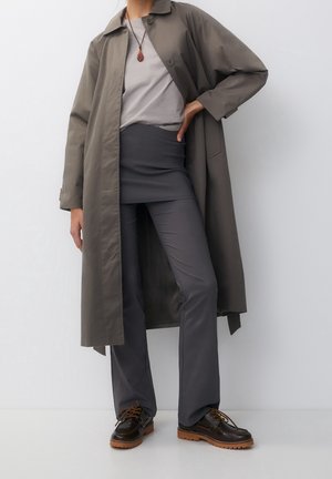 Osoba ubrana w szary płaszcz trencz, jasnoszarą koszulę, ciemnoszare spodnie z nakładką spódnicową oraz ciemnobrązowe buty z grubymi podeszwami, stojąca przy białej ścianie.