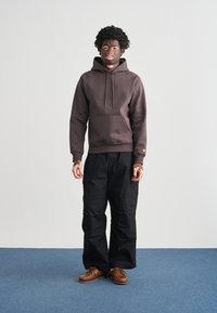 Σκούφο σκούρου καφέ χρώματος με μπροστινή τσέπη, σε συνδυασμό με μαύρο παντελόνι εργασίας με πολλές τσέπες. Το μοντέλο φοράει καφέ παπούτσια. Απλό φόντο.