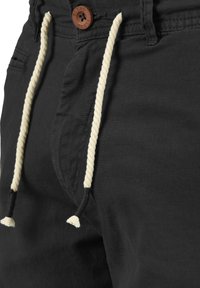 Pantalones cortos negros con una tela texturizada, con un botón marrón y un cordón beige con borlas. Primer plano del área de la cintura.