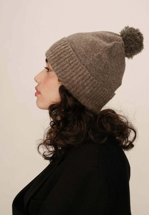 Bonnet en tricot marron avec une texture côtelée, doté d'un pompon duveteux sur le dessus. Porté sur une tête avec des cheveux noirs et bouclés.