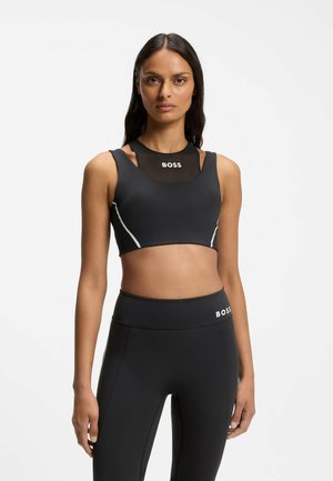 Kvinde iført sort atletisk crop top og leggings med BOSS-logo, stående mod en ensfarvet hvid baggrund.