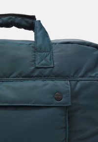Geantă de culoare teal închis, cu un design matlasat, având un mâner superior, un buzunar frontal cu buton de tip snap și detalii de cusătură contrastante.