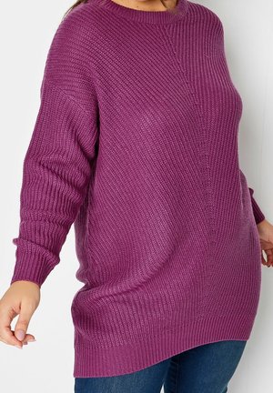 Nainen, joka pukeutunut teksturoituun violettiin ylisuureen neulepuseroon, jonka hihoissa on pitkät hihansuut ja resorit, yhdistettynä sinisiin farkkuihin, yksiväristä taustaa vasten.